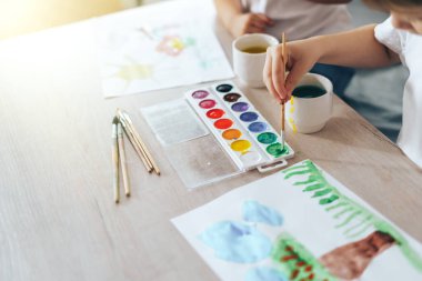 Children paint green grass with a tree and blue clouds.