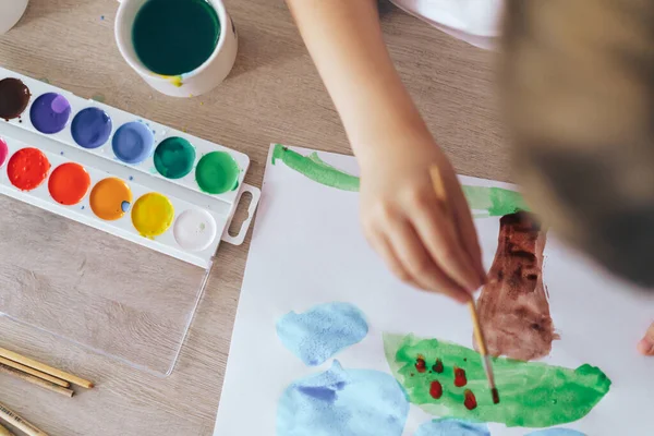 Children paint green grass with a tree and blue clouds.