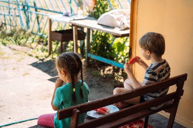 Children sitting on a bench eat watermelon.