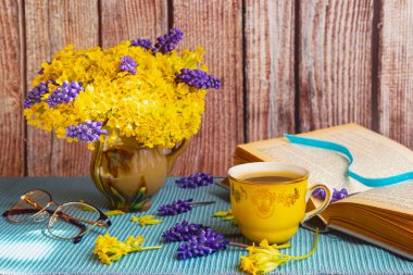 Vazoda sarı çuha çiçekleri ve muscari var. Ahşap arka plan, kitap, sarı bardak, gözlük. Mavi masa örtüsü. Yan görünüm.