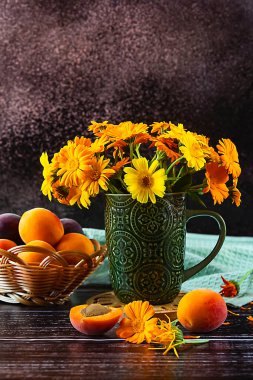 Yellow calendula flowers in a green cup on the table. Apricots in a basket. Composition on the table on a dark background. Side view.