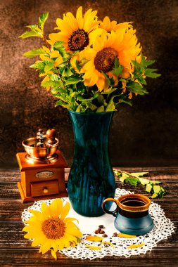 A bouquet of sunflowers in a blue vase on a dark background in a rustic style. Blue cup of coffee and vintage coffee grinder on the table. Side view