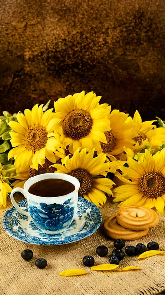 A bouquet of sunflowers on a dark background in a rustic style. A white and blue cup, blueberries and cookies on the table. Summer still life.Top view