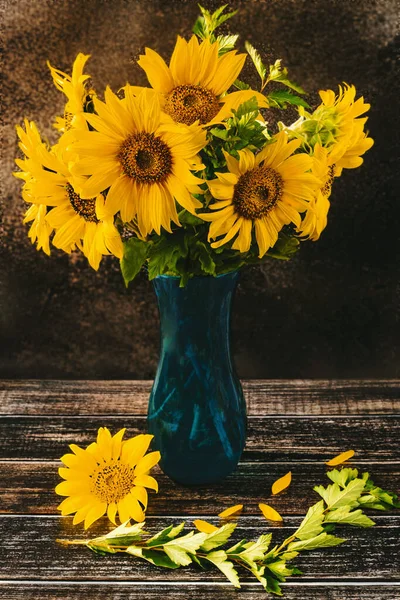 A bouquet of sunflowers in a blue vase on a dark background in a rustic style. Summer still life, side view.