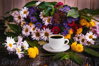 Beyaz bir fincan kahve ve ahşap arka planda renkli bir buket sonbahar çiçeği. Yan görünüm.