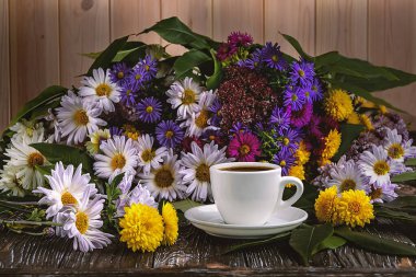 Beyaz bir fincan kahve ve ahşap arka planda renkli bir buket sonbahar çiçeği. Yan görünüm.