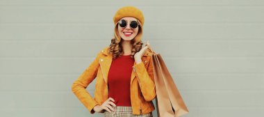 Gri arka planda beresi olan, sarı ceketli, güzel, mutlu, alışveriş torbalarıyla gülümseyen genç bir kadının sonbahar portresi.