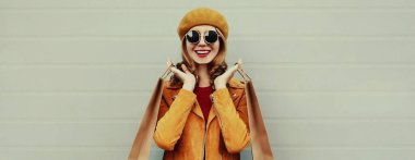 Gri arka planda beresi olan, sarı ceketli, güzel, mutlu, alışveriş torbalarıyla gülümseyen genç bir kadının sonbahar portresi.