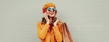 Alışveriş çantasıyla gülen genç bir kadının sonbahar portresi. Akıllı telefondan arıyor. Sarı ceketli, gri arka planda bere takıyor.