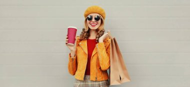 Güz portresi, güzel, mutlu, gülümseyen, alışveriş torbaları ve kahve fincanı olan, sarı ceketli, gri arka planda bere takan genç bir kadın.