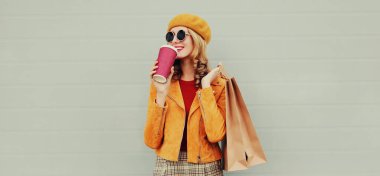 Güz portresi, güzel, mutlu, gülümseyen, alışveriş torbaları ve kahve fincanı olan, sarı ceketli, gri arka planda bere takan genç bir kadın.