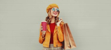 Güz portresi, güzel, mutlu, gülümseyen, alışveriş torbaları ve kahve fincanı olan, sarı ceketli, gri arka planda bere takan genç bir kadın.