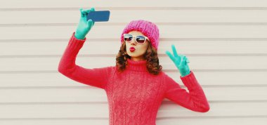 Akıllı telefonuyla selfie çeken, örgü şapkalı, beyaz arka planda süveter giyen genç bir kadının portresi.