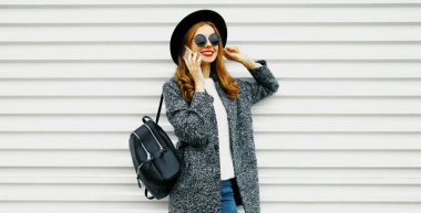 Mutlu gülümseyen sarışın bir kadının portresi. Beyaz arka planda gri bir palto, yuvarlak bir şapka ve sırt çantası takan bir akıllı telefondan arıyor.