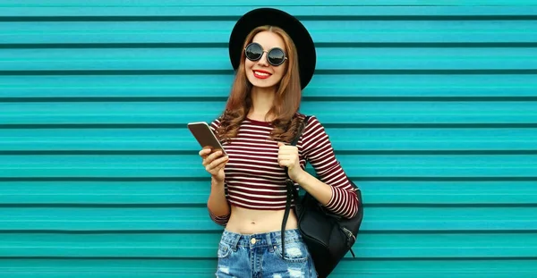 青い背景に帽子をかぶっているスマートフォンを持つ幸せな笑顔の若い女性の肖像画 — ストック写真