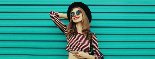 青い背景に帽子をかぶっているスマートフォンを持つ幸せな笑顔の若い女性の肖像画 — ストック写真