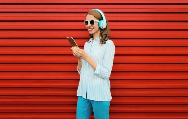 Kulaklıklı gülümseyen genç bir kadının portresi kırmızı arka planda akıllı telefonuyla müzik dinliyor.