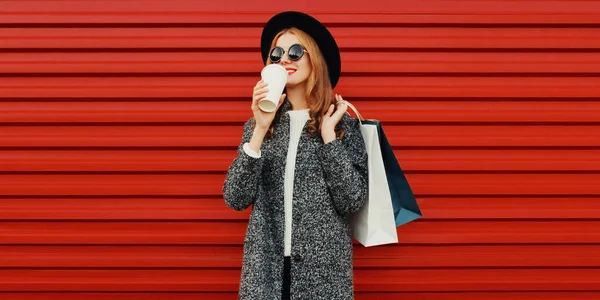 グレーのコートを着てショッピングバッグと美しい笑顔の女性の肖像画 カラフルな赤の背景に帽子 — ストック写真