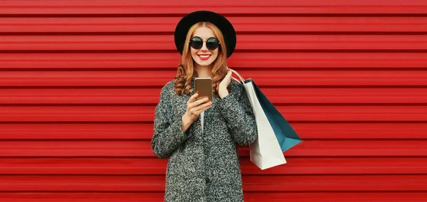ショッピングバッグやグレーのコートを着たスマートフォンを持つ幸せな笑顔の女性の肖像画 カラフルな赤い背景の帽子 — ストック写真