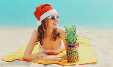 Kırmızı Noel Baba şapkalı mutlu genç bir kadının Noel portresi. Mavi deniz arka planında birlikte uzanan komik ananaslı.