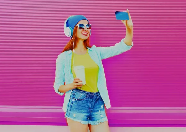 ピンクを背景に音楽を聴くヘッドフォンでスマートフォンで自画撮りをするスタイリッシュな幸せな笑顔の若い女性の肖像 — ストック写真