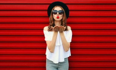 Güzel genç bir kadının portresi dudaklarını silip kırmızı arka planda siyah bir şapka takarak hava öpücüğü yolluyor.