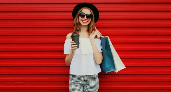 赤い背景に黒丸帽子をかぶってコーヒーのカップとショッピングバッグと美しい笑顔の女性の肖像画 — ストック写真