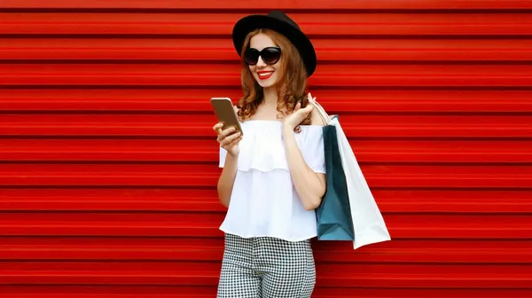 色彩斑斓的红色背景下 带着购物袋和智能手机的快乐微笑的女人的画像 — 图库照片