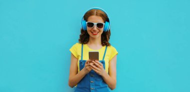 Kulaklıklı gülümseyen genç bir kadının portresi mavi arka planda akıllı telefonuyla müzik dinliyor.