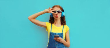 Moda portresi kulaklıklı genç bir kadının mavi arka planda akıllı telefonuyla müzik dinlemesi.
