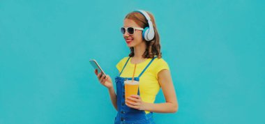 Kulaklıklı gülümseyen genç bir kadının portresi mavi arka planda akıllı telefonuyla müzik dinliyor.