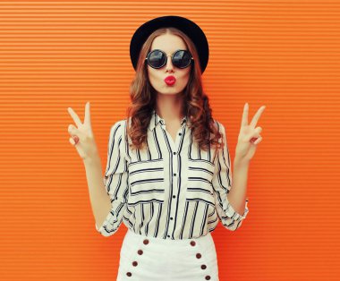 Güzel genç bir kadının portresi dudaklarını silip beyaz çizgili bir gömlek ve turuncu arka planda siyah bir şapka giyerek hava öpücüğü yolluyor.