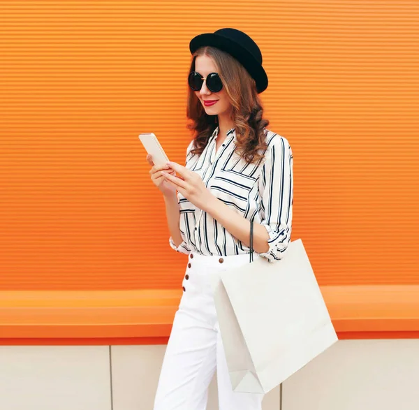 Alışveriş çantasıyla telefona bakan, beyaz çizgili gömlek giyen, turuncu arka planda siyah şapkalı güzel bir genç kadının portresi.