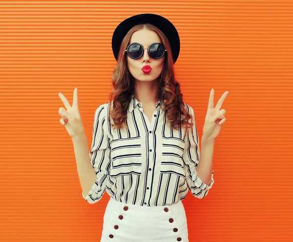 Güzel genç bir kadının portresi dudaklarını silip beyaz çizgili bir gömlek ve turuncu arka planda siyah bir şapka giyerek hava öpücüğü yolluyor.