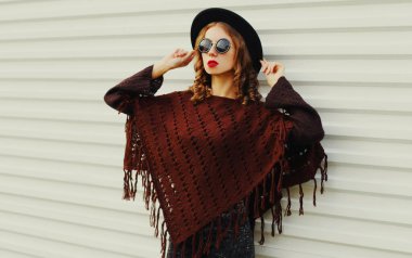 Kahverengi örgü panço giyen güzel bir genç kadın portresi, beyaz arka planda siyah yuvarlak şapka.