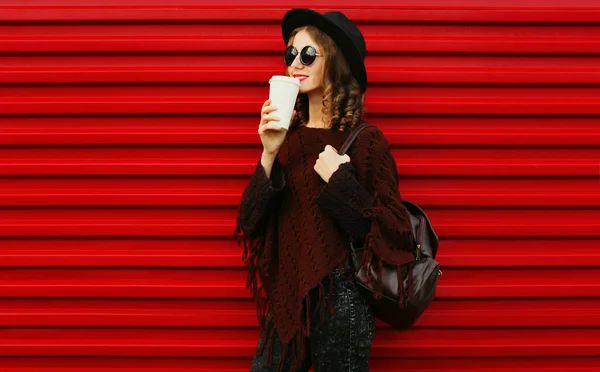 茶色いニットのポンチョを着たコーヒーを飲みながら美しい若い女性の肖像 赤い背景に黒い丸い帽子 — ストック写真