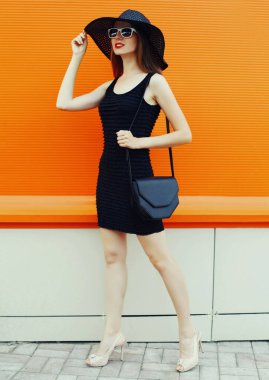 El çantalı güzel bir kadının portresi. Siyah bir şapka ve turuncu arka planda elbise giyiyor.