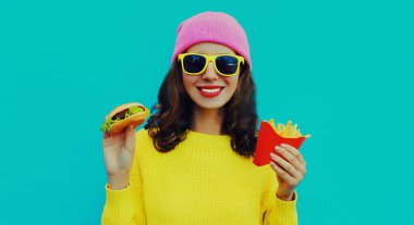 Modaya uygun gülümseyen genç bir kadının portresi hamburger ve patates kızartması ile mavi renkli arka planda.
