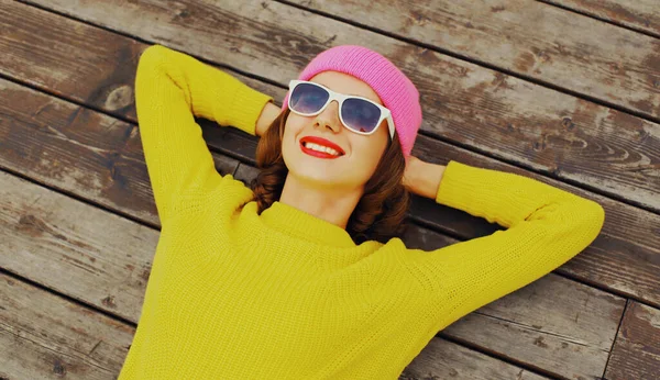 黄色のニットセーターとピンクの帽子を身に着けているスタイリッシュな笑顔の若い女の子の肖像画 ティーネージャーは 市公園内の木製の床に横たわってリラックス — ストック写真