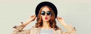 Tarz sahibi bir kadının portresi dudaklarını sümkürerek tatlı bir hava öpücüğü gönderiyor gri arka planda siyah yuvarlak bir şapka takıyor.