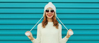 Mutlu gülümseyen genç bir kadının kışın portresi beyaz örgü bir süveter, mavi arka planda ponponlu bir şapka giyerek eğleniyor.