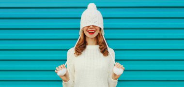 Mutlu gülümseyen genç bir kadının kışın portresi beyaz örgü bir süveter, mavi arka planda ponponlu bir şapka giyerek eğleniyor.