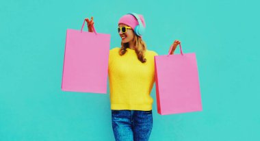 Kulaklıkla müzik dinleyen, sarı örgü süveter, mavi arka planda pembe şapka giymiş, kulaklık takan, şık bir genç kadının renkli portresi.