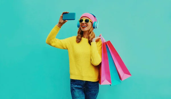 黄色のニットセーター ピンクの帽子を身に着けているショッピングバッグとヘッドフォンで電話で自撮りを取るスタイリッシュな幸せな笑顔の女性青の背景 — ストック写真