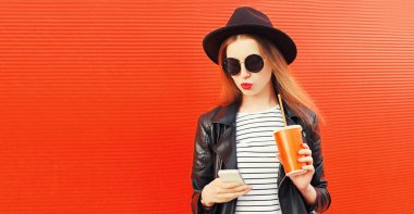 Şık bir genç kadın portresi, akıllı telefon ve bir bardak meyve suyu ile siyah ceketli, kırmızı arka planda şapka takıyor.