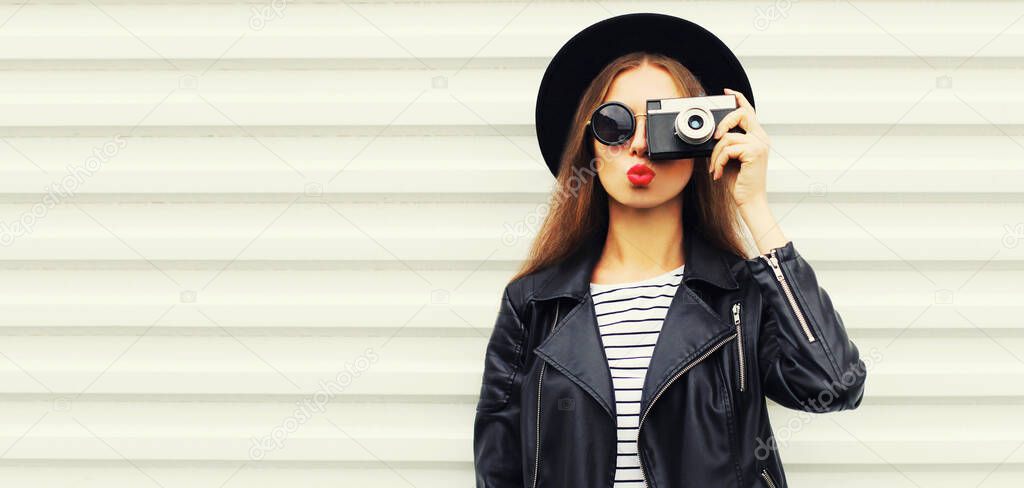 Stylish portrait of happy young woman photographer with vintage film camera wearing a black round hat, leather jacket on white background