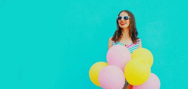 Mavi arka planda renkli bir elbise giyen ve bir sürü balonu olan mutlu, güzel bir genç kadının portresi.