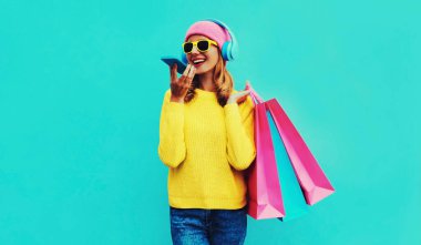 Şık, gülümseyen genç bir kadının renkli portresi. Alışveriş çantalarıyla arıyor kulaklıkla müzik dinliyor sarı örgü bir kazak giyiyor, mavi arka planda pembe şapka takıyor.