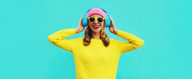 Mavi arka planda renkli sarı süveter giyen, kulaklıklı, mutlu gülümseyen genç bir kadının portresi.