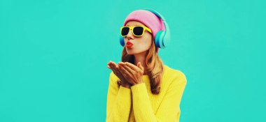 Kulaklıkla müzik dinleyen şık bir genç kadının portresi mavi arka planda renkli sarı bir kazak giyerek tatlı bir hava öpücüğü gönderiyor.